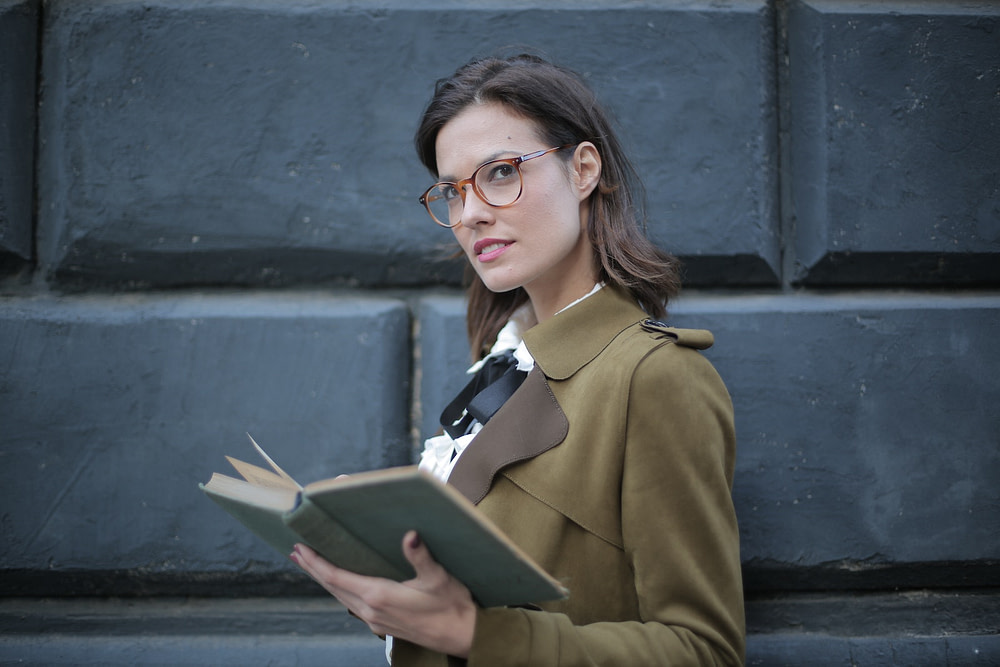 From below confident young female in glasses and elegant vintage clothes looking away and contemplating while reading rook on street near black wall of aged building in city